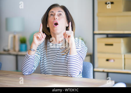Mittleren Alters ältere Frau am Tisch sitzen zu Hause erstaunt und überrascht mit der Suche und zeigt mit Fingern und erhobenen Armen. Stockfoto