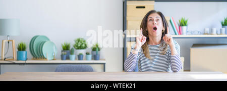 Weitwinkel Foto von mittleren Alters ältere Frau am Tisch sitzen zu Hause erstaunt und überrascht suchen und zeigt mit Fingern und erhobenen Armen. Stockfoto