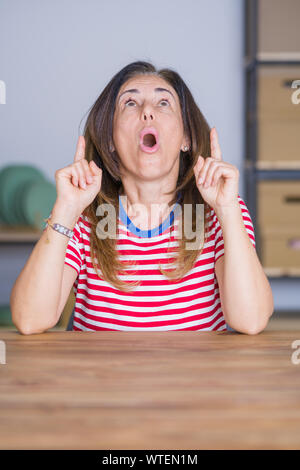 Mittleren Alters ältere Frau am Tisch sitzen zu Hause erstaunt und überrascht mit der Suche und zeigt mit Fingern und erhobenen Armen. Stockfoto