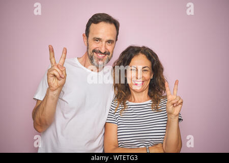 Schönen mittleren Alter Paar zusammen über isolierte rosa Hintergrund stand lächelnd mit glücklichen Gesicht in die Kamera winking tun Sieg unterzeichnen. Anzahl t Stockfoto