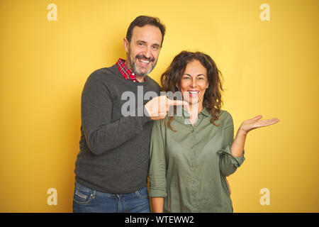 Schönen mittleren Alter Paar über isolierte gelben Hintergrund erstaunt und lächelt in die Kamera während der Präsentation mit Hand und zeigte mit dem Finger. Stockfoto