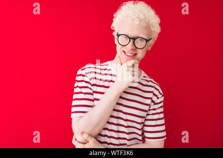 Junge albino blonder Mann tragen gestreifte T-Shirt und Brille über isolierten roten Hintergrund in die Kamera lächelt zuversichtlich, mit verschränkten Armen und Stockfoto