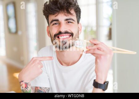 Junger Mann essen asiatische Sushi mit Stäbchen mit Überraschung Gesicht zeigenden Finger zu sich selbst Stockfoto
