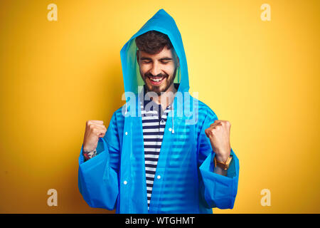 Jungen gutaussehenden Mann tragen Regenjacke mit Kapuze über isolierte gelben Hintergrund sehr glücklich und aufgeregt, Sieger Geste mit erhobenen Armen, Stockfoto