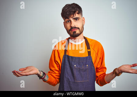 Junge Ladenbesitzer Mann mit Tattoo tragen Schürze stehend über isoliert weißer Hintergrund ratlos und verwirrt Ausdruck mit den Armen und hob die Hand. Tun Stockfoto