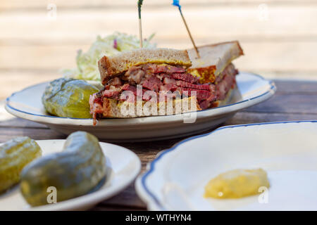 Pastrami Sandwich in der Hälfte auf Roggenbrot in Scheiben geschnitten mit Senf auf einer weißen Platte auf einem Holz Tisch von oben gesehen. Krautsalat und Dill Pickles an der Seite. Stockfoto