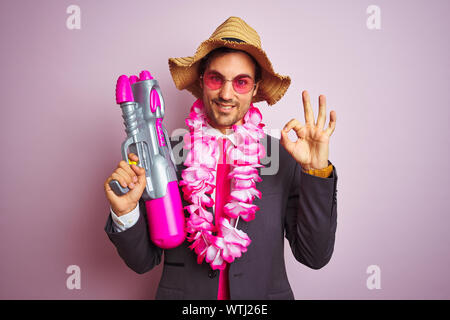 Junge Unternehmer tragen Wasserpistole Hawaii Lei Hut Brille über isolierte rosa Hintergrund tun ok Zeichen mit den Fingern, ausgezeichnete Symbol Stockfoto