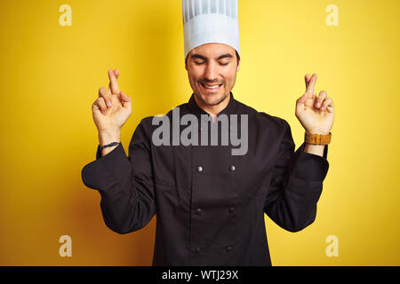 Junge Küchenchef Mann in Uniform und Mütze stehend über isolierte gelben Hintergrund und deutete mit dem Finger lächelnd mit Hoffnung und überquert die Augen geschlossen. Glück und s Stockfoto