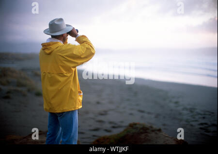 Ansicht von hinten von einem Mann durch ein Fernglas auf einem ruhigen Strand suchen. Stockfoto