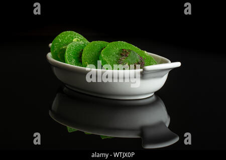 Gruppe von vier Scheiben süß grün kandierte Kiwis in weiße, ovale Keramik Schüssel auf schwarzem Glas isoliert Stockfoto