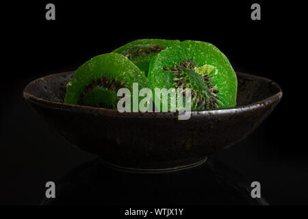 Gruppe von drei Scheiben süß grün kandierte Kiwis in dunklen Keramik Schüssel auf schwarzem Glas isoliert Stockfoto