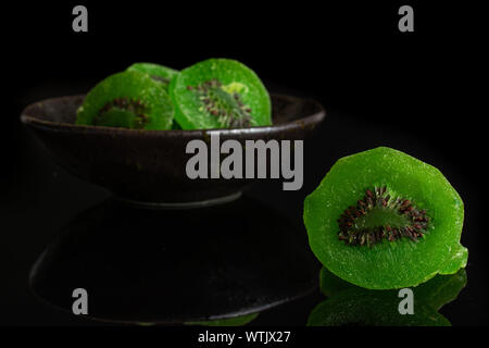 Gruppe von vier Scheiben süß grün kandierte Kiwis in dunklen Keramik Schüssel auf schwarzem Glas isoliert Stockfoto