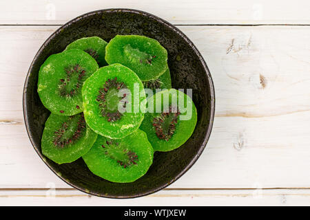 Viele Scheiben süß grün kandierte Kiwis in dunklen Keramik Schüssel flatlay auf weißem Holz Stockfoto