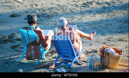 Alicante, Spanien, 25. August 2019: Zwei ältere Frauen am Strand Kreuzworträtsel lösen und lesen Sie beim Bräunen sitzt. Sommer für Rentner oder Pensionäre. Stockfoto