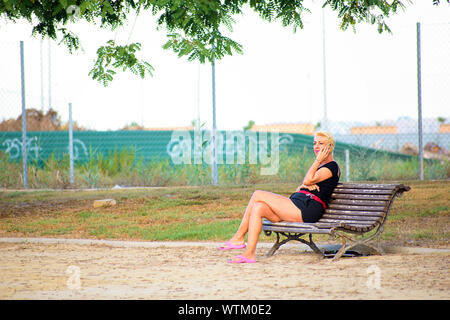 Eine glückliche einsame Frau oder ein Mädchen sitzen, in einer Bank im Park im Sommer lächelnd warten auf jemanden und über Ihr Telefon sitzt, Murcia Stockfoto