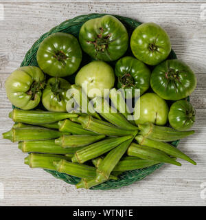Frische organische grüne Tomaten und Okra in einem Korb auf einem weißen Holz board isoliert. Stockfoto