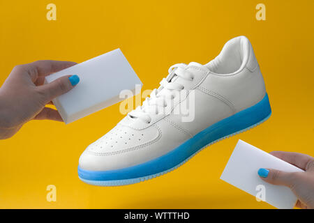 Trendige weisse Turnschuhe mit blauer Sohle und weibliche Hände mit Schwämmen auf gelbem Hintergrund, Schuhe Care Concept Stockfoto