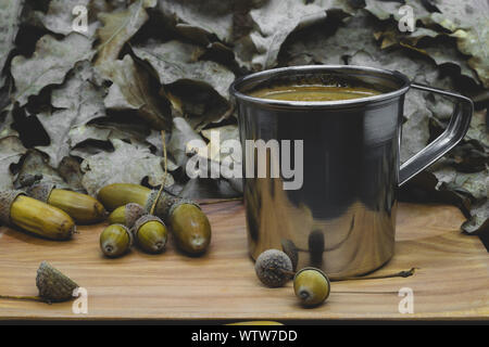 Abstrakte Kompositionen aus gekochten Acorn weiß Kaffee in Metall Becher und Eiche Eicheln auf Holzplatte im Hintergrund der Eiche Blätter Stockfoto