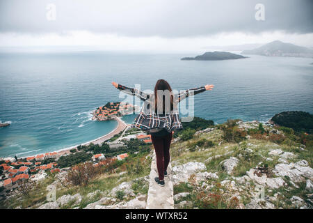 Das Mädchen hebt die Hände hoch. Sie bewundert die schöne Aussicht auf die Insel Sveti Stefan in Montenegro. Stockfoto