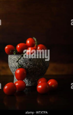 Kleine, helle rote Tomaten auf dem Zweig Schuß in einem grauen Stein Schüssel mit Holz Hintergrund. Früchte sind in der Tabelle zum Ausdruck. Natürliches Licht.
