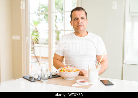 Mittleres Alter mann Essen asiatisch Essen mit Stäbchen zu Hause mit ernsten Gesichtsausdruck. Einfache und natürliche in die Kamera schaut. Stockfoto
