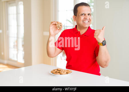 Mittleres Alter Mann essen Chocolate Chips cookies zu Hause zeigen und zeigt mit dem Daumen bis zu der Seite mit glücklichen Gesicht lächelnd Stockfoto