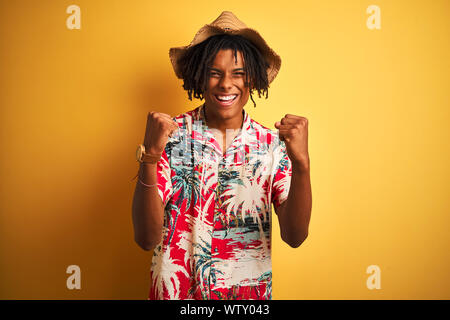 Afro-amerikanische Mann mit Dreadlocks tragen Blumen Shirt und Hut über isolierte gelben Hintergrund feiern überrascht und für den Erfolg mit Waffen erstaunt Stockfoto