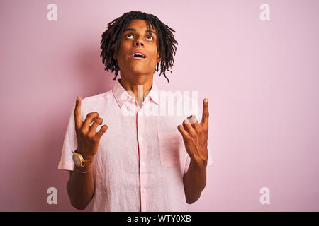 Afro-amerikanische Mann mit Dreadlocks tragen elegante Hemd stehend über isolierte rosa Hintergrund erstaunt und überrascht mit der Suche und zeigt mit finge Stockfoto
