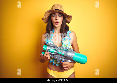 Schöne Frau das Tragen von Bikinis und Lei hawaiianischen Blumen und Wasser Gewehr über isolierte Hintergrund im Schlag mit einer Überraschung Gesicht, Angst und Excite Angst Stockfoto