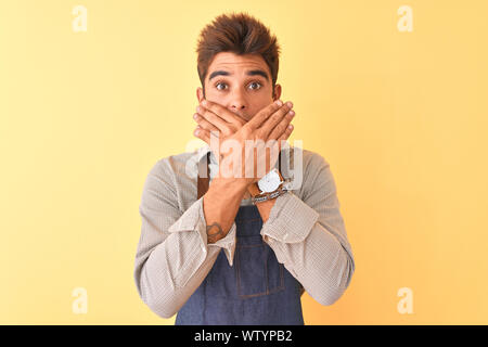Jungen gutaussehenden Mitarbeiter Mann mit Schürze stehend über isolierte gelben Hintergrund schockiert über den Mund mit Händen für Fehler. Geheime Konzept. Stockfoto
