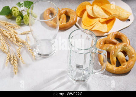 Sauber und leer Bier Glas und eine Tasse mit verschiedenen Snacks rund - Brezeln und Chips. Bier Zutaten - Hopfen und Gerste in den Hintergrund. Stockfoto