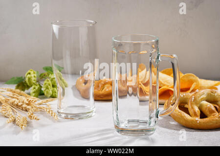 Sauber und leer Bier Glas und eine Tasse mit verschiedenen Snacks rund - Brezeln und Chips. Bier Zutaten - Hopfen und Gerste in den Hintergrund. Stockfoto