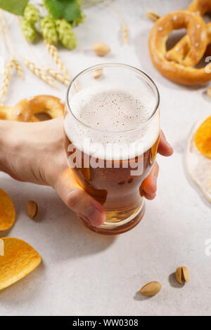 Mann hand mit einem Glas helles Bier. Verschiedene Snacks im Hintergrund - Brezeln, Chips und Nüsse. Stockfoto