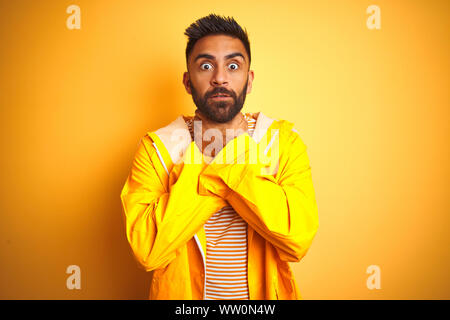 Junge indische Mann mit regenmantel stehend über isolierte gelben Hintergrund schreien und Ersticken, weil schmerzhaft ersticken. Gesundheitliches Problem. Aufheben Stockfoto