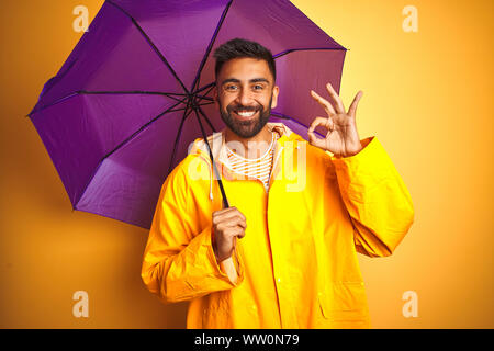 Junge indische Mann mit Regenmantel und lila Schirm über isolierte gelben Hintergrund tun ok Zeichen mit den Fingern, ausgezeichnete Symbol Stockfoto