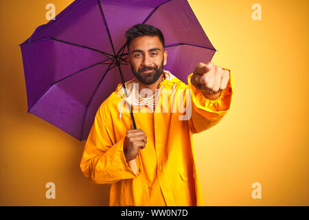 Junge indische Mann mit Regenmantel und lila Schirm über isolierte gelber Hintergrund mit dem Finger an die Kamera und an Sie gerichtet, Handzeichen, positi Stockfoto