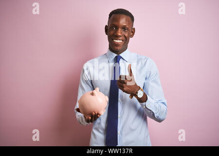 Afrikanische amerikanische Geschäftsmann holding Sparschwein stehend über isolierte rosa Hintergrund zufrieden mit Lächeln tun ok Zeichen, Daumen hoch, mit den Fingern, PE Stockfoto