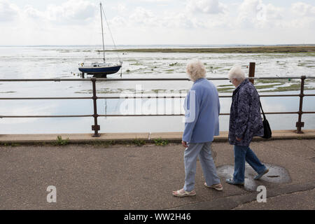 Zwei ältere Damen zu Fuß entlang der Promenade, wo eine einzelne Yacht sitzt aufrecht in Ebbe Mündung Schlamm an alten Leigh, am 10. September 2019, in Leigh-on-Sea, Essex, England. Stockfoto