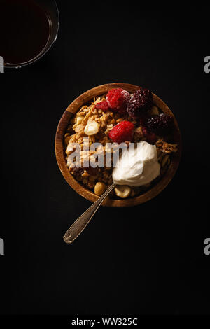 Müsli mit Himbeeren und Brombeeren und einen Löffel Sahne in Houten mit einer Tasse Kaffee auf Schwarz. Stockfoto