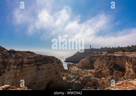 Felsen, Hügel und Täler und felsige Landschaft an der Küste der Algarve mit, in der Ferne, die Alfanzina Leuchtturm, Portugal Stockfoto