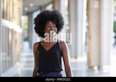 Lächelnde junge schwarze Frau zu Fuß in der Stadt genießen Sie den sonnigen Tag mit geschlossenen Augen Stockfoto