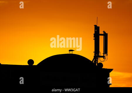 Ein Mobiltelefon Sender auf dem Dach eines Gebäudes in Leeds, gegen eine orange sonnendurchfluteten Himmel Silhouette Stockfoto