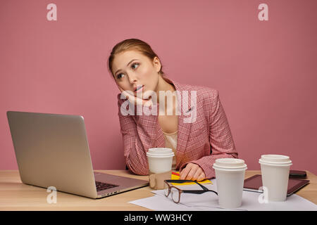 Rothaarige Mädchen im Anzug auf einem rosa Hintergrund sitzt vor einem Laptop und müde unterstützt ihren Kopf mit der Hand. Stockfoto