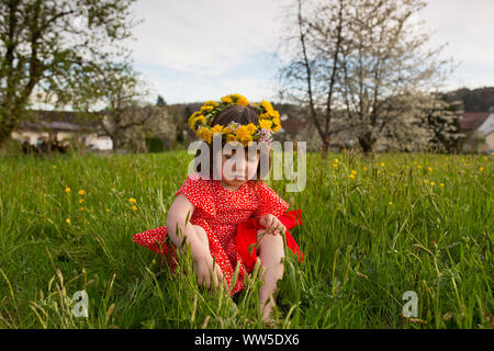 5-jähriges Mädchen im roten Kleid und Blume Kranz sitzen in einer grünen Wiese Stockfoto