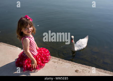 4-6 Jahre alten Mädchen in der rot gestreiften Kleid sitzt am Ufer, Schwan im Wasser Stockfoto