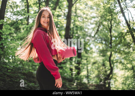 7/8 Bild einer attraktiven Athlet Frau mit flatternden Haare in ein sportliches Sweatshirt mit Kapuze mit einem Sport Matte auf einem Waldweg. Stockfoto
