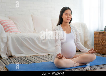 Das Meditieren auf Mutterschaft. Nahaufnahme des Asiatischen jungen schwangeren Frau meditieren Während in Lotus Position zu Hause sitzen. Stockfoto