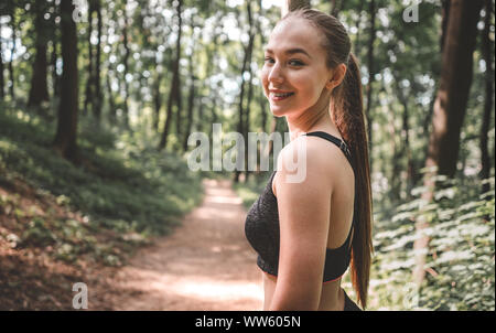 Portrait einer jungen sportliche Frau mit einem Lächeln auf ihrem Gesicht auf in den Wald. Schöne Mädchen suchen und lächelnd in die Kamera vor dem Ausführen Stockfoto