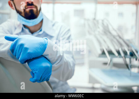 Attraktive stattliche Zahnarzt in schützende Maske, weißen Kittel auf dem Stuhl sitzt. Zahnarzt Ausrüstungen für den Hintergrund. Konzentrieren Sie sich auf die Hände. Stockfoto