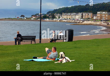 Ein paar Leute sitzen und entspannen am Meer, Urlaub Stadt, von Largs auf den Firth of Clyde in Schottland. Es ist ein Mann, die auf dem Gras mit einer ziemlich großen, schwarzen und weißen Hund sein Auge auf die Dinge! Alan Wylie/ALAMY © Stockfoto